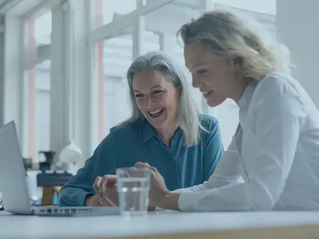 Zwei lächelnde Frauen, die im Büro an einem Schreibtisch sitzen und gemeinsam an einem Laptop arbeiten.